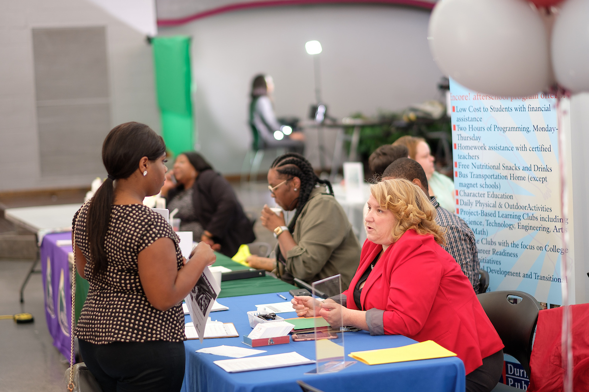 A woman in a red blazer speaks with an attendee at an information fair.