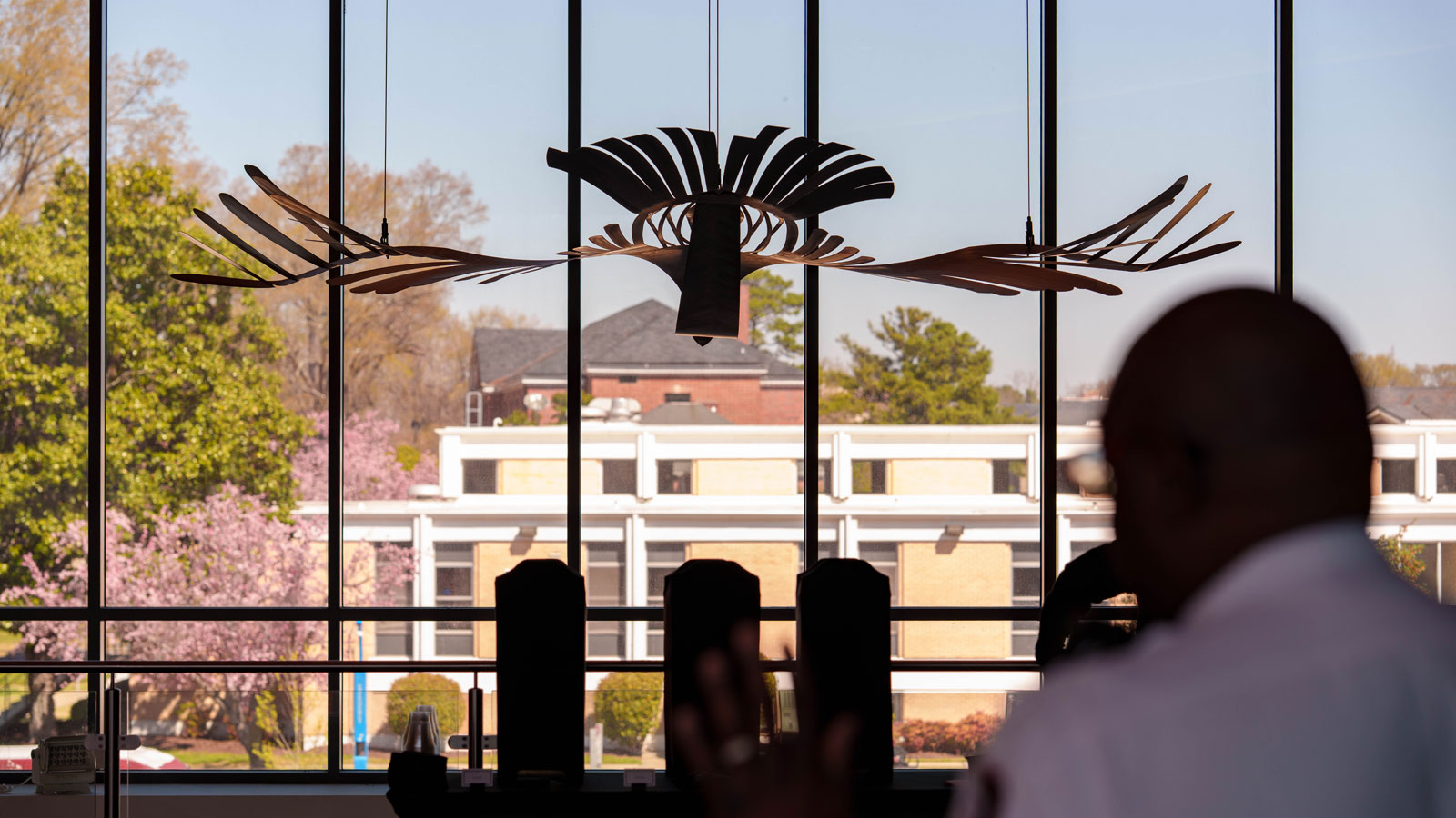 A dark, bird-like sculpture hangs in front of a large window, overlooking buildings and trees. A blurred figure with their hand raised is in the foreground.