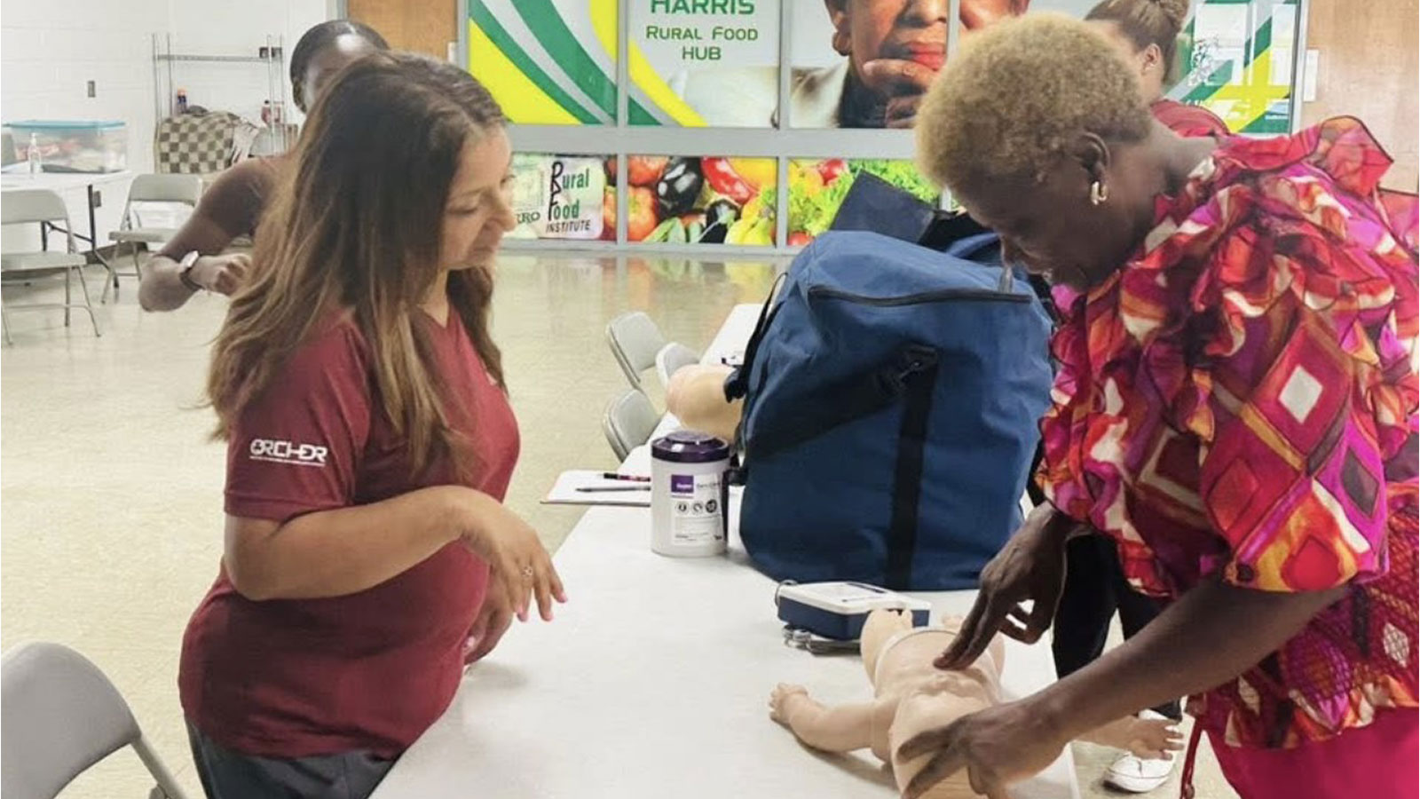 A woman in a red shirt teaches another woman, in a colorful shirt, how to perform CPR on a baby mannequin.