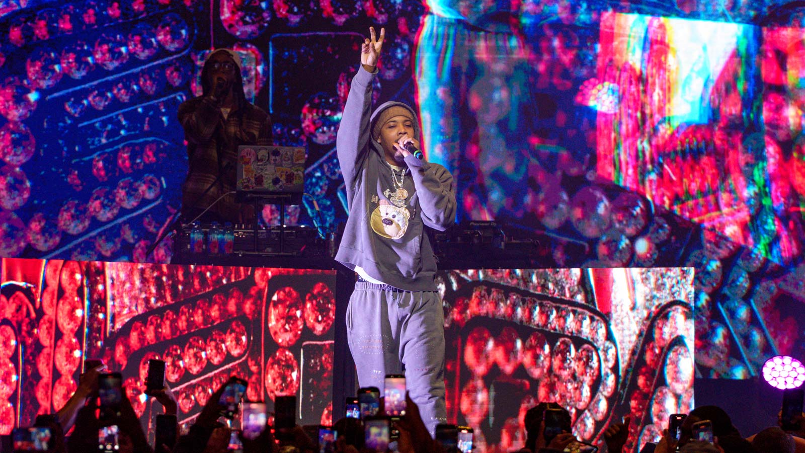 Rapper G Herbo holds up a peace sign while performing on stage in front of a crowd holding up smartphones.