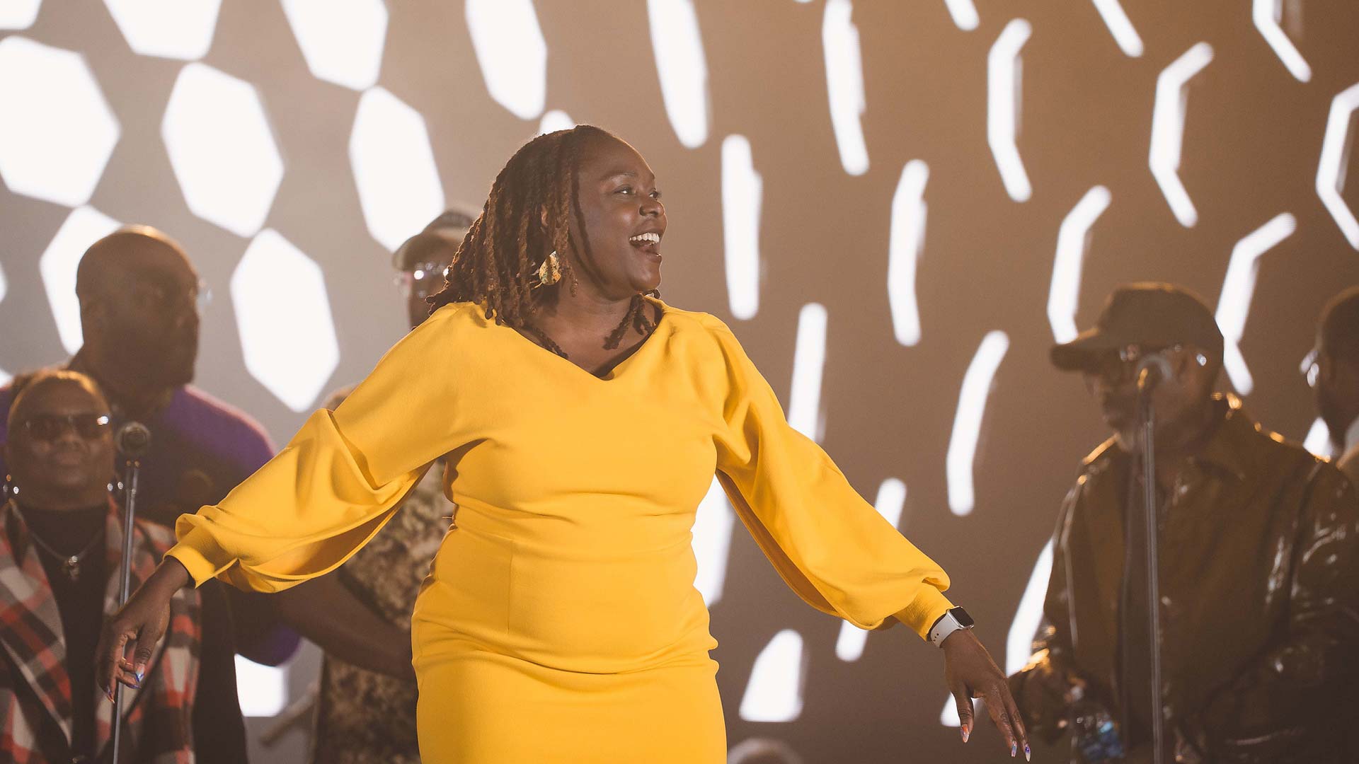 A smiling woman in a long-sleeved bright yellow dress sings onstage in front of a choir with a patterned backdrop illuminated behind them.