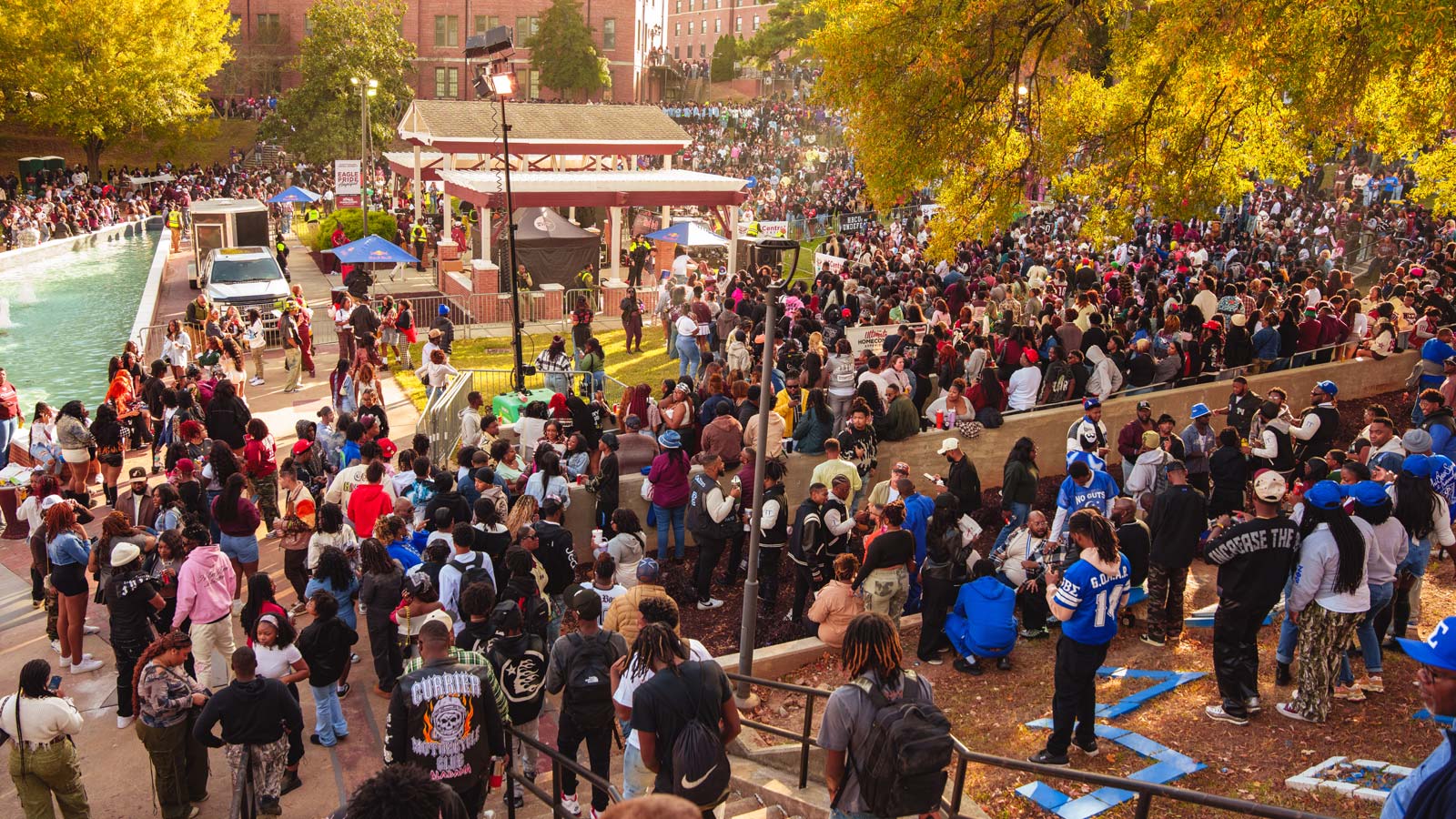 A large crowd gathers around a campus courtyard and reflecting pool surrounded by autumn trees during a university event.