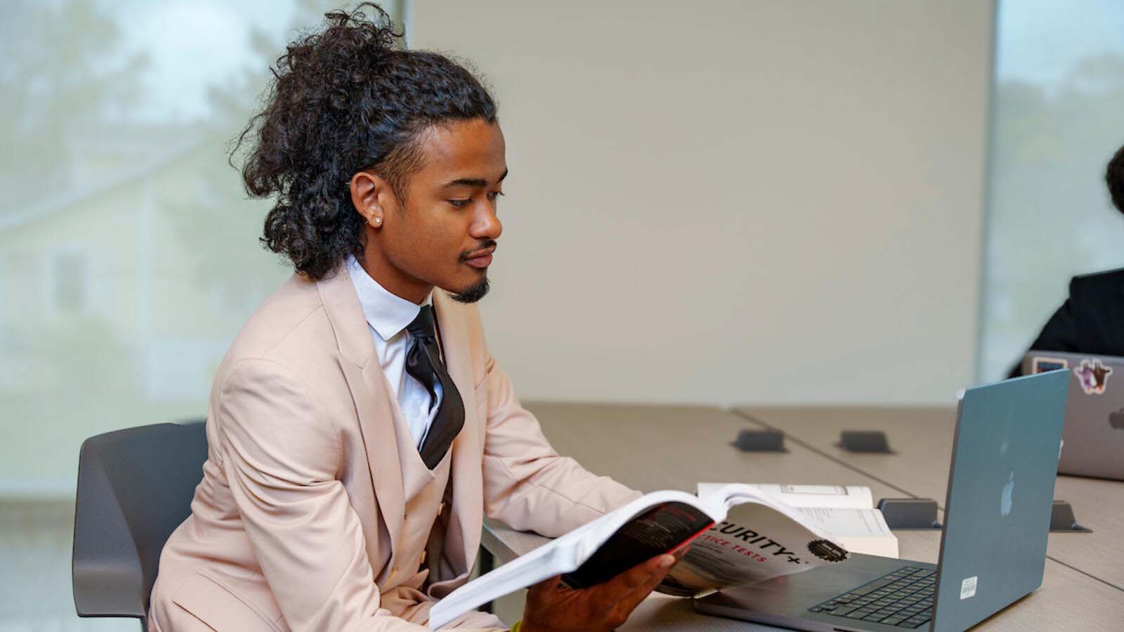 A student in a light beige suit reads a textbook while working at a laptop inside a brightly lit study space.
