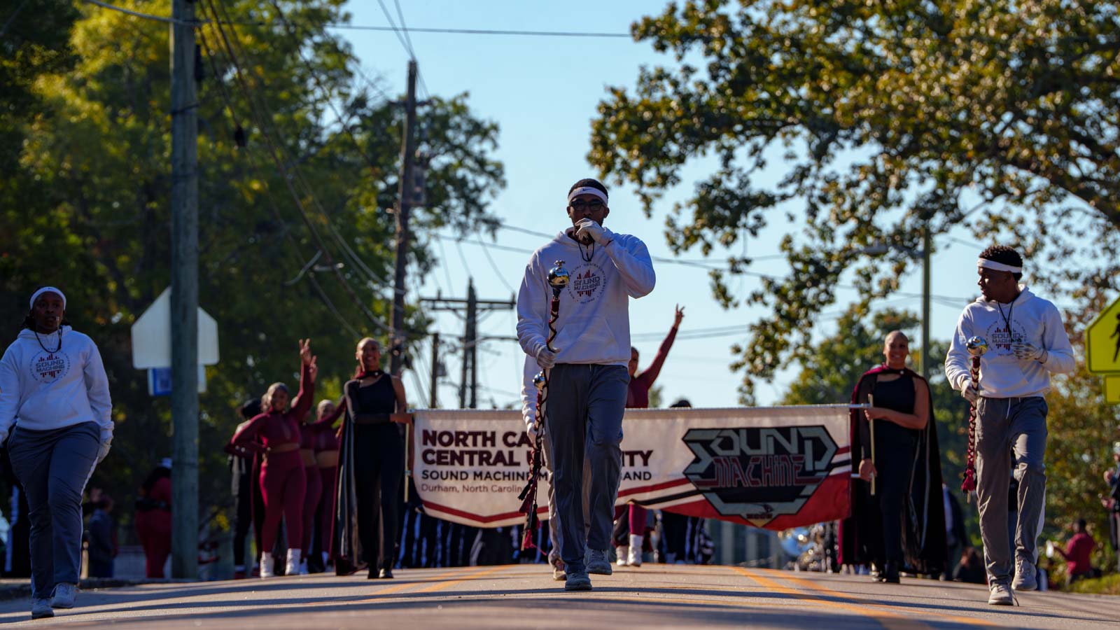 Drum majors and band members march down a street behind a banner for the North Carolina Central University Sound Machine Marching Band.