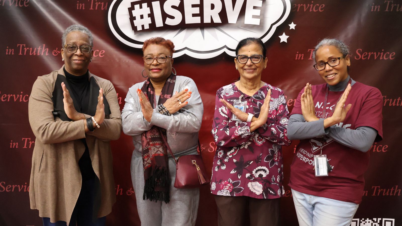 Four women pose with crossed arms in a service gesture during a North Carolina Central University community engagement event, standing in front of an #ISERVE backdrop.