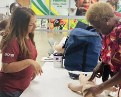 A woman in a red shirt teaches another woman, in a colorful shirt, how to perform CPR on a baby mannequin.