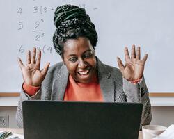  A smiling woman with her hands raised is looking at a laptop, with math equations visible on a whiteboard behind her, suggesting a virtual teaching or learning environment.