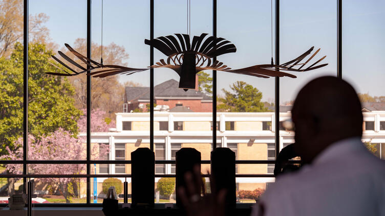 A dark, bird-like sculpture hangs in front of a large window, overlooking buildings and trees. A blurred figure with their hand raised is in the foreground.