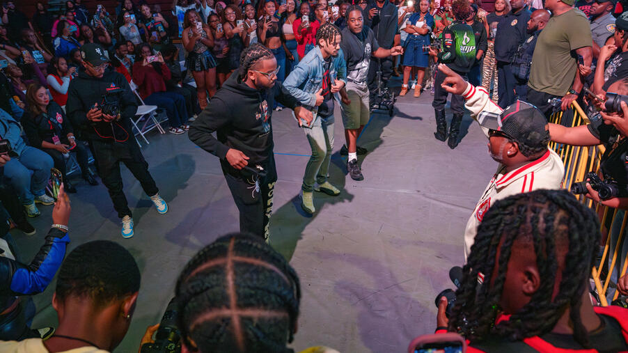 A group of young men perform in the center of an open arena floor surrounded by a crowd recording with their phones.
