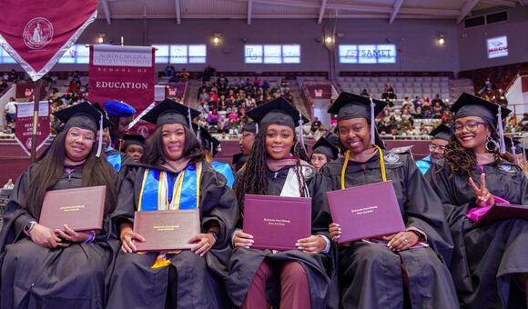 Graduates in caps and gowns holding diplomas, smiling at a ceremony.