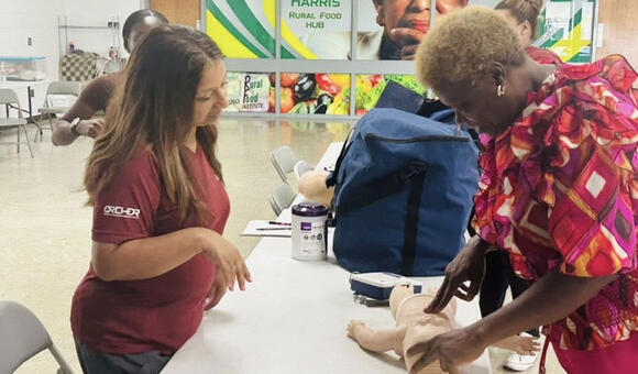 A woman in a red shirt teaches another woman, in a colorful shirt, how to perform CPR on a baby mannequin.