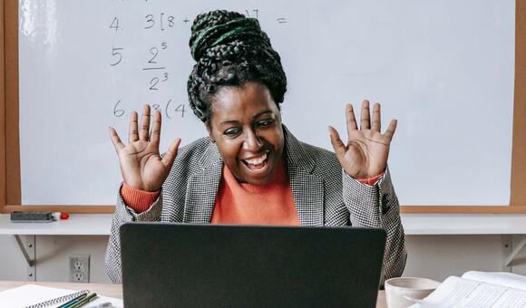  A smiling woman with her hands raised is looking at a laptop, with math equations visible on a whiteboard behind her, suggesting a virtual teaching or learning environment.