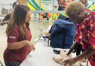  A woman in a red shirt teaches another woman, in a colorful shirt, how to perform CPR on a baby mannequin.