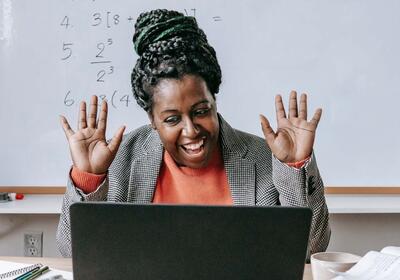  A smiling woman with her hands raised is looking at a laptop, with math equations visible on a whiteboard behind her, suggesting a virtual teaching or learning environment.